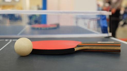 Nahaufnahme eines Tischtennisschlägers mit Ball auf einem Tischtennistisch mit Netz im Hintergrund.