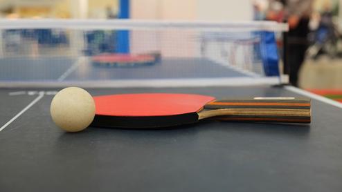 Nahaufnahme eines Tischtennisschlägers mit Ball auf einem Tischtennistisch mit Netz im Hintergrund.