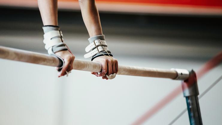 Nahaufnahme zweier Hände, die eine schmale Turnstange mit Griffpolstern halten, im Hintergrund eine Turnhalle.